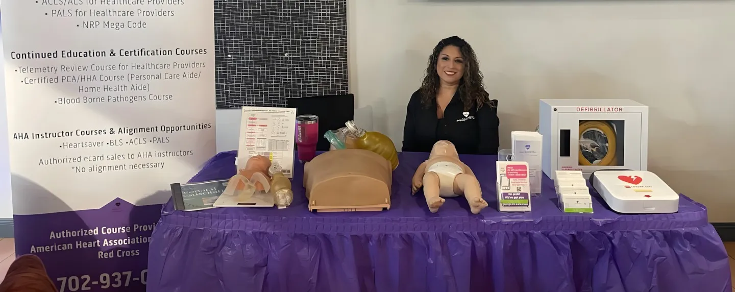 Adriana Park, RN, at her Las Vegas CPR training table with infant and adult manikins, a bag-valve mask, AHA materials, and an AED