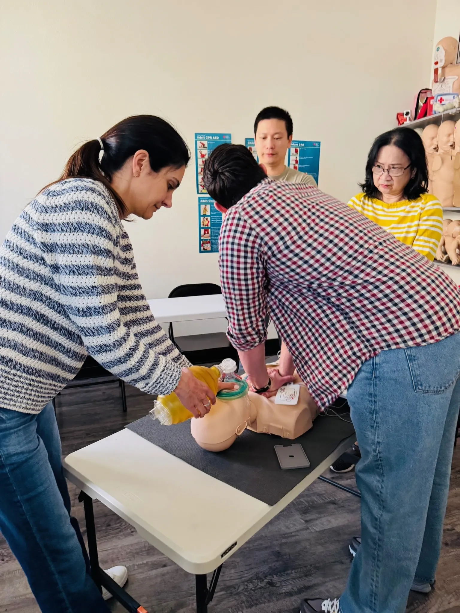 Three students at a Heartbeat Haven training table — center student bagging an adult CPR manikin with a bag-valve-mask while two classmates assist, AHA CPR posters visible on the wall behind