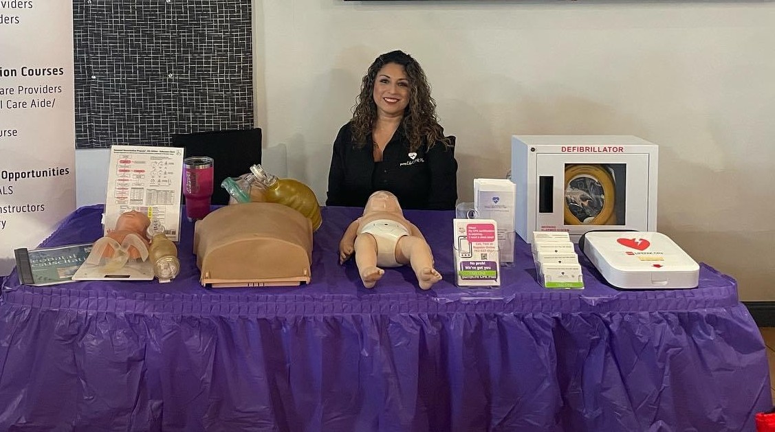 Adriana Park, MSN, RN at a Heartbeat Haven CPR demo table with adult and infant manikins, a bag-valve-mask, an AHA reference card, and a defibrillator